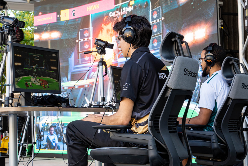 Two headphoned young people sit in roller chairs on stage where their game of Super Smash Bros is being projected on to a screen behind them.