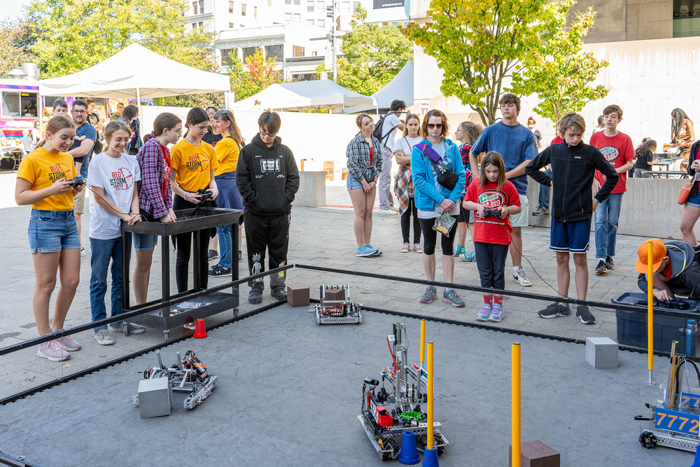 Young people gather round a closed-off square of pavement where they operate remote controlled robots.