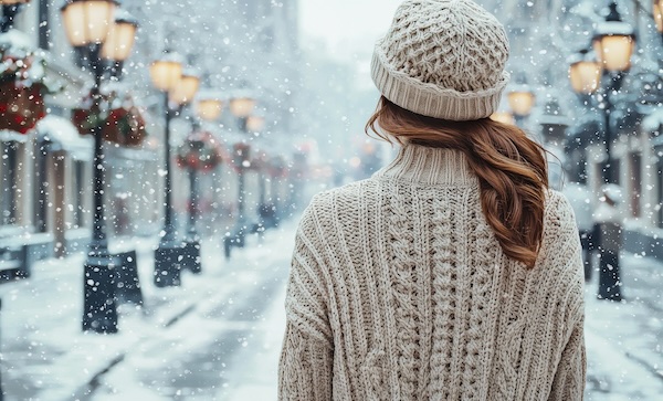 Vintage woman in cozy knit sweater walking down snow-covered city street with winter decorations
